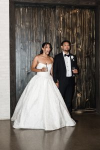 A bride in a strapless white gown and a groom in a black tuxedo stand side by side, holding hands and drinks, looking ahead with anticipation, captured by a Toronto wedding photographer against a dark wooden backdrop.