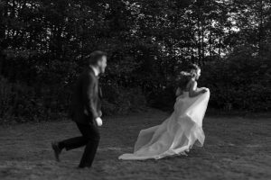 A black and white documentary-style photo captures a bride in a flowing dress running across grass, with a man in a suit chasing her. Trees fill the background, evoking a candid moment of joy in Toronto photography.