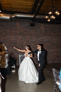 A bride in a white gown and a groom in a tuxedo, both wearing sunglasses, dance playfully together at a wedding reception with brick walls and industrial lighting, capturing an editorial vibe perfect for creative wedding photography.