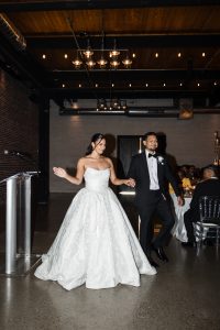 A bride in a white strapless gown and a groom in a black tuxedo smile as they enter a modern, industrial-style wedding venue with exposed brick walls and hanging lights, captured by a documentary photographer.