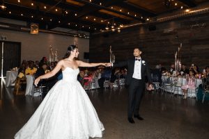A bride in a white gown and a groom in a black tuxedo dance together in a warmly lit Toronto wedding reception hall, as guests seated at decorated tables watch and smile in the background—a perfect moment captured through photography.
