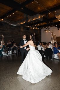 A bride in a white strapless gown and a groom in a black tuxedo dance together in a warmly lit reception hall, as seated guests watch—a beautiful moment captured with an editorial photography style.