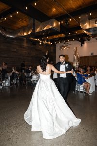 A bride in a white gown and a groom in a tuxedo smile at each other while dancing in a warmly lit reception hall, surrounded by seated guests watching them—a perfect moment captured by a Toronto documentary photographer.