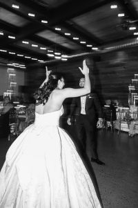 A wedding photographer captures a bride in a strapless gown facing her groom as they dance joyfully in a stylish, dimly lit reception hall filled with seated guests.
