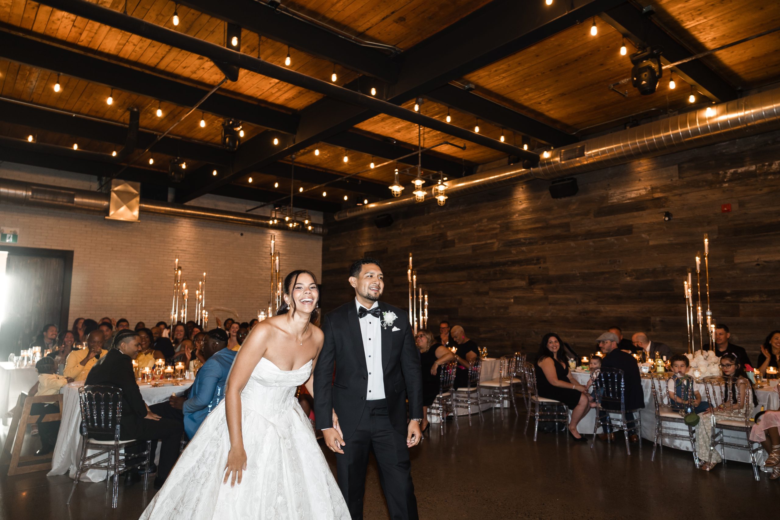 A bride in a strapless white gown and a groom in a black tuxedo smile and hold hands in a warmly lit reception hall, as a skilled photographer captures this documentary moment with guests seated at decorated tables in the background.