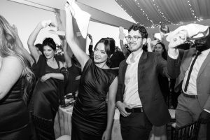 A group of well-dressed wedding guests at a formal event stand and wave napkins in the air, smiling and celebrating under a tent decorated with string lights—perfect for an editorial photographer to capture.