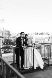 A bride in a strapless gown leans on the shoulder of a groom in a tuxedo as they stand together on a rooftop balcony, looking out over a cityscape. This black and white wedding photography captures a candid, documentary moment.