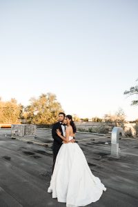 A bride in a white gown and a groom in a black tuxedo stand together on a rooftop, embracing under a clear sky with trees in the background—captured by an editorial photographer in Toronto.