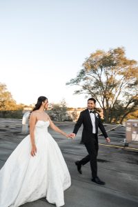 A bride in a white wedding dress and a groom in a black tuxedo hold hands and walk outdoors, smiling at each other, as a Toronto photographer captures their moment with trees and a clear sky in the background.