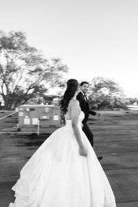 A bride in a white gown and a groom in a suit walk hand in hand outdoors, smiling at each other. Captured by a documentary photographer, the black and white photo features blurred trees and industrial equipment in the background.