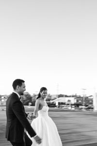 A black and white editorial photo of a bride and groom holding hands and smiling as they walk outdoors, with a blurred cityscape in the background—capturing the timeless essence of wedding photography.