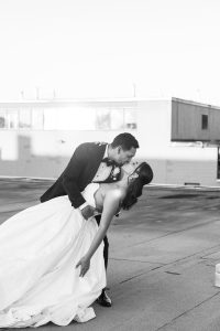A groom in a tuxedo dips and kisses his bride, who is wearing a strapless wedding dress, on a rooftop. This editorial wedding moment is captured in stunning black and white.