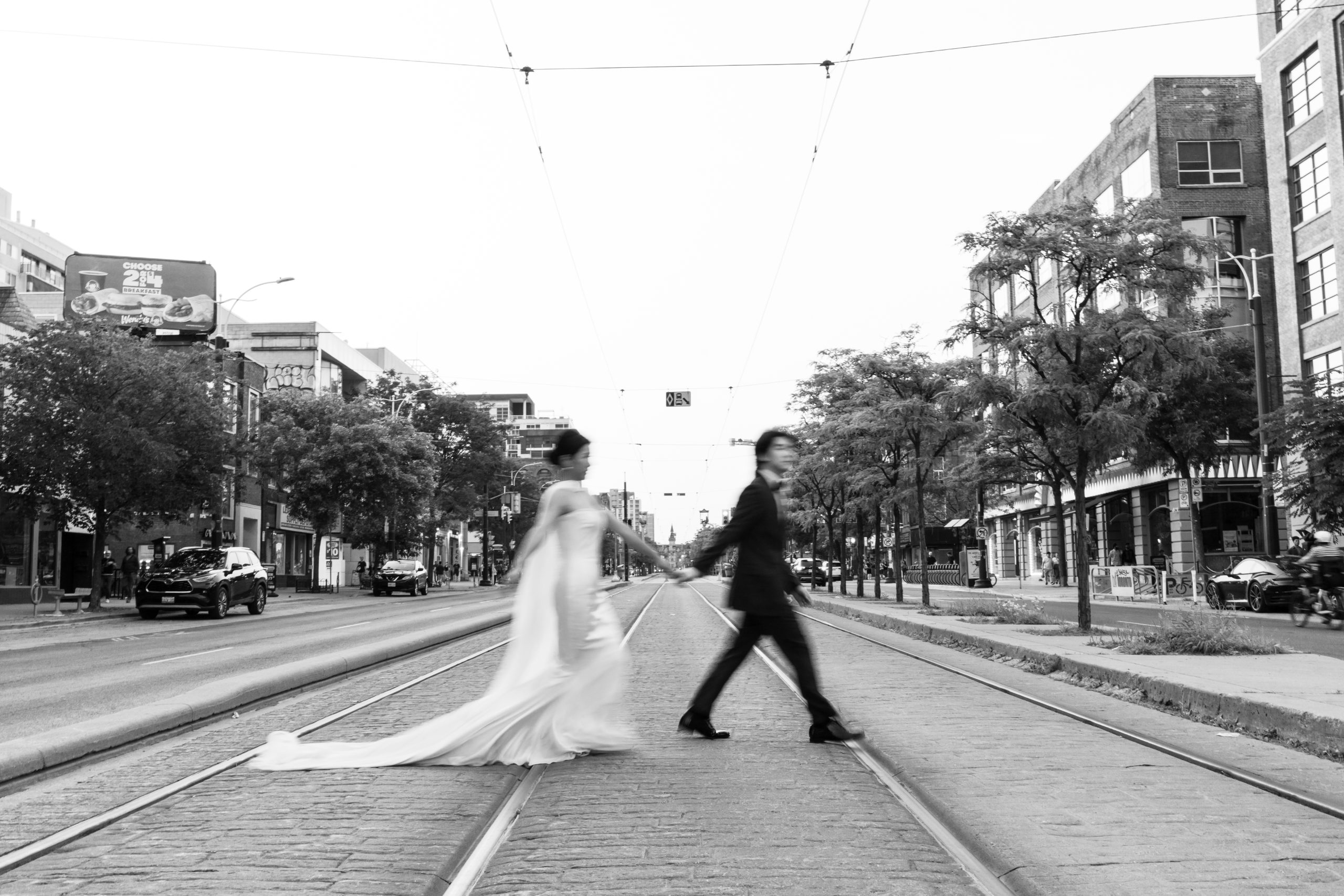 A bride and groom walk hand in hand across Toronto streetcar tracks in a city, captured in black and white with motion blur, surrounded by buildings and trees—a timeless documentary wedding moment.