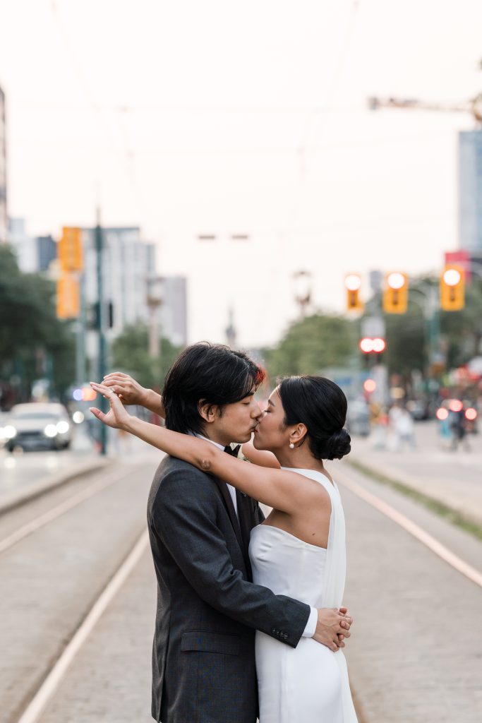 A couple dressed in wedding attire kisses on a city street in Toronto, standing on tram tracks with arms wrapped around each other. Buildings, cars, and traffic lights blur into the background, capturing a perfect wedding photography moment.