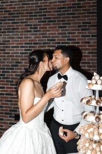 A bride and groom in wedding attire share a kiss while holding cannoli, standing next to a tiered display of pastries, with a brick wall in the background—a perfect moment for a documentary photographer’s editorial portfolio.