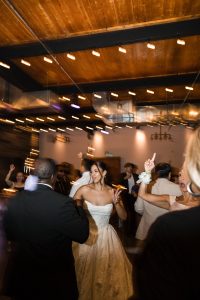A bride in a white dress dances and smiles joyfully at a lively indoor wedding reception, surrounded by guests under warm string lights and a wooden ceiling, capturing the essence of wedding photography.