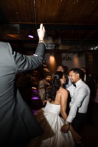 A bride in a white strapless gown and a groom in a white shirt and bow tie laugh and dance closely together at a lively indoor wedding reception, captured in an editorial style amid the joyful crowd of guests.