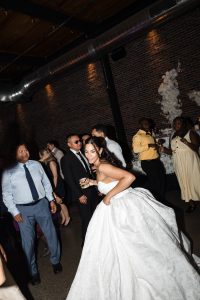 A bride in a white gown dances at a wedding reception, smiling and holding a drink, as guests in formal attire gather in an industrial-style Toronto venue with brick walls—captured beautifully by expert photography.