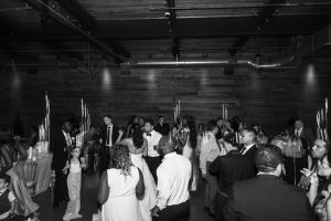 A black-and-white documentary photo captures people dressed formally, dancing and mingling at a lively indoor event, possibly a wedding reception, with candlelit tables in the background and a wooden wall.