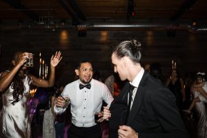 Two men in formal attire, one in a white shirt and bow tie, the other in a black suit, hold drinks and chat while dancing at a lively Toronto wedding. Other elegantly dressed guests are seen dancing in the background, adding to the documentary feel.