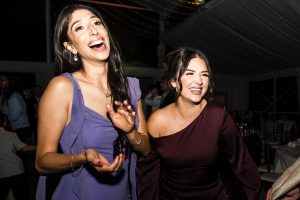 Two women in formal dresses laugh and smile together at a lively indoor event, captured by a documentary photographer, with string lights and guests visible in the background.
