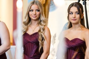 Two women in matching strapless burgundy dresses stand side by side, looking in different directions, as a Toronto wedding photographer captures them with soft lighting and blurred white objects in the foreground.