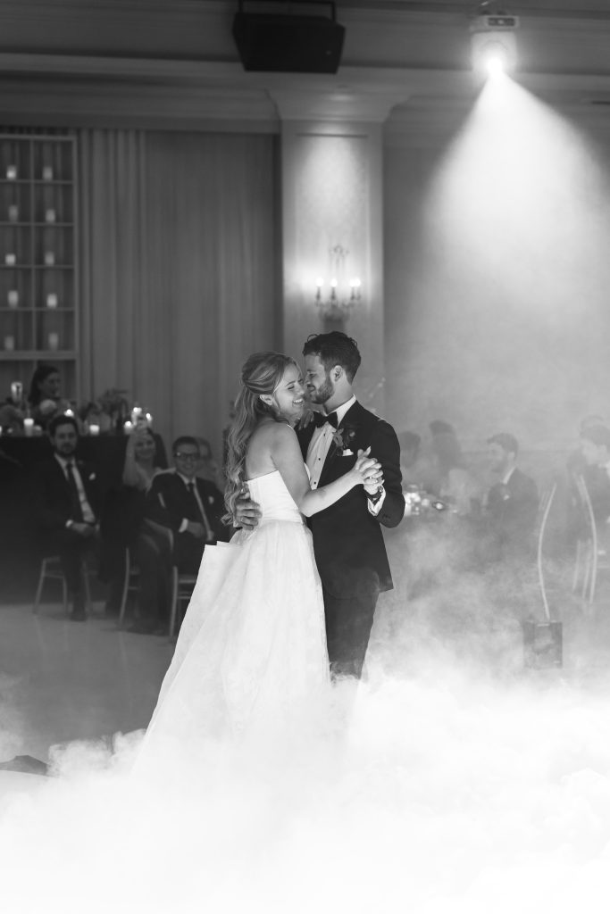 A bride and groom share their first dance in a romantic, fog-filled Toronto ballroom. Captured by a skilled wedding photographer, the bride glows in her strapless gown while the groom looks sharp in his tuxedo, surrounded by delighted guests.