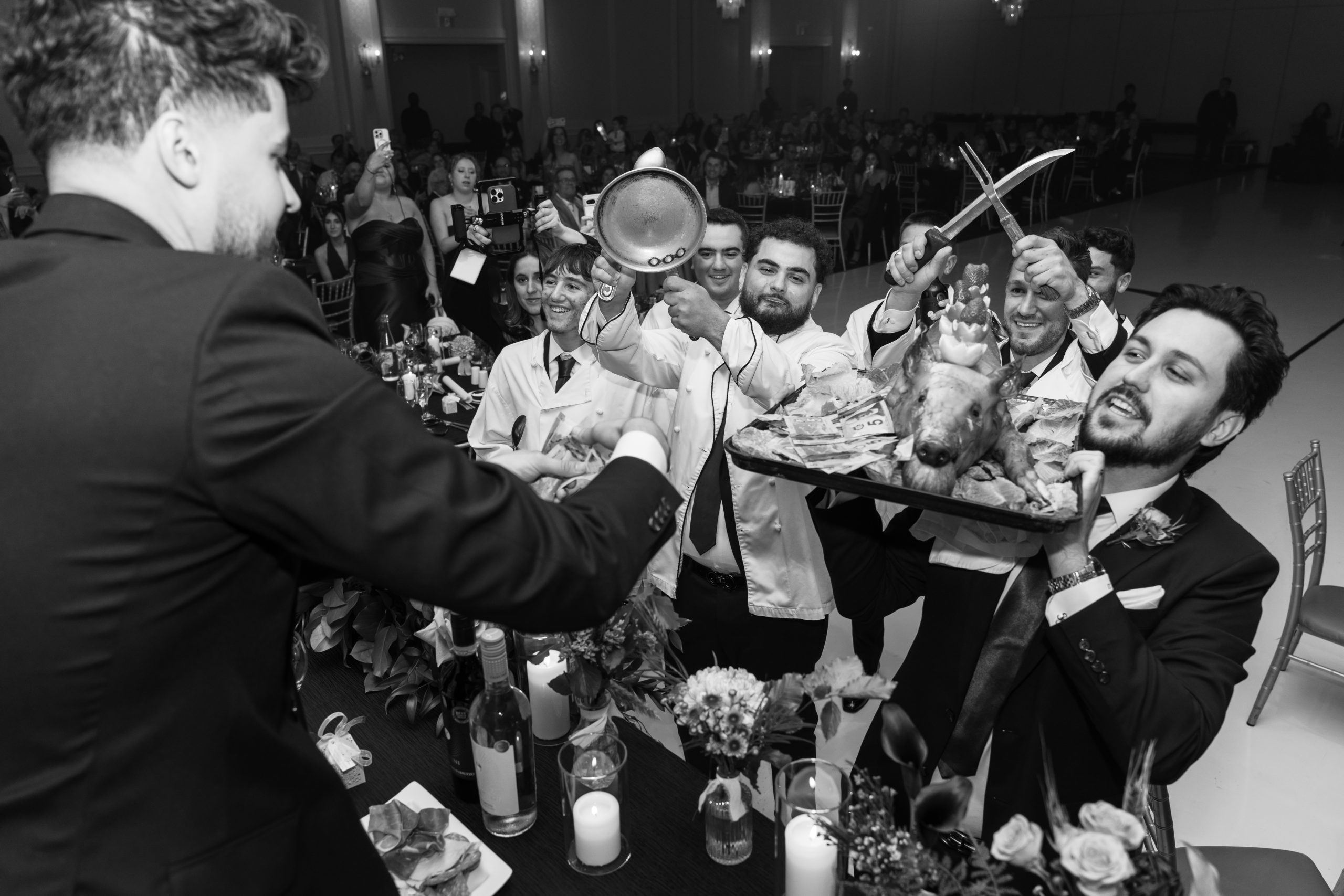 A group of people, including chefs holding kitchen utensils, present a large platter with a cooked animal’s head and garnishes to a man in a suit at a formal Toronto wedding, as onlookers and a photographer capture the moment among decorated tables.