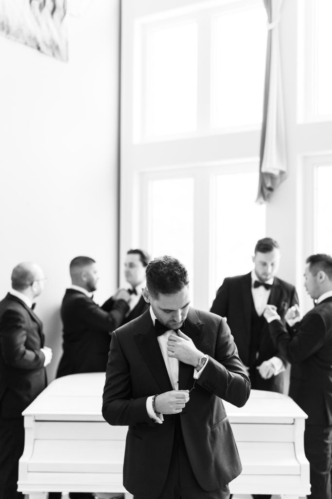 A group of men in tuxedos stand by a white piano in a bright, sunlit room. As one adjusts his cufflink, others talk and get ready in the background—a perfect moment of wedding photography captured by a skilled photographer.