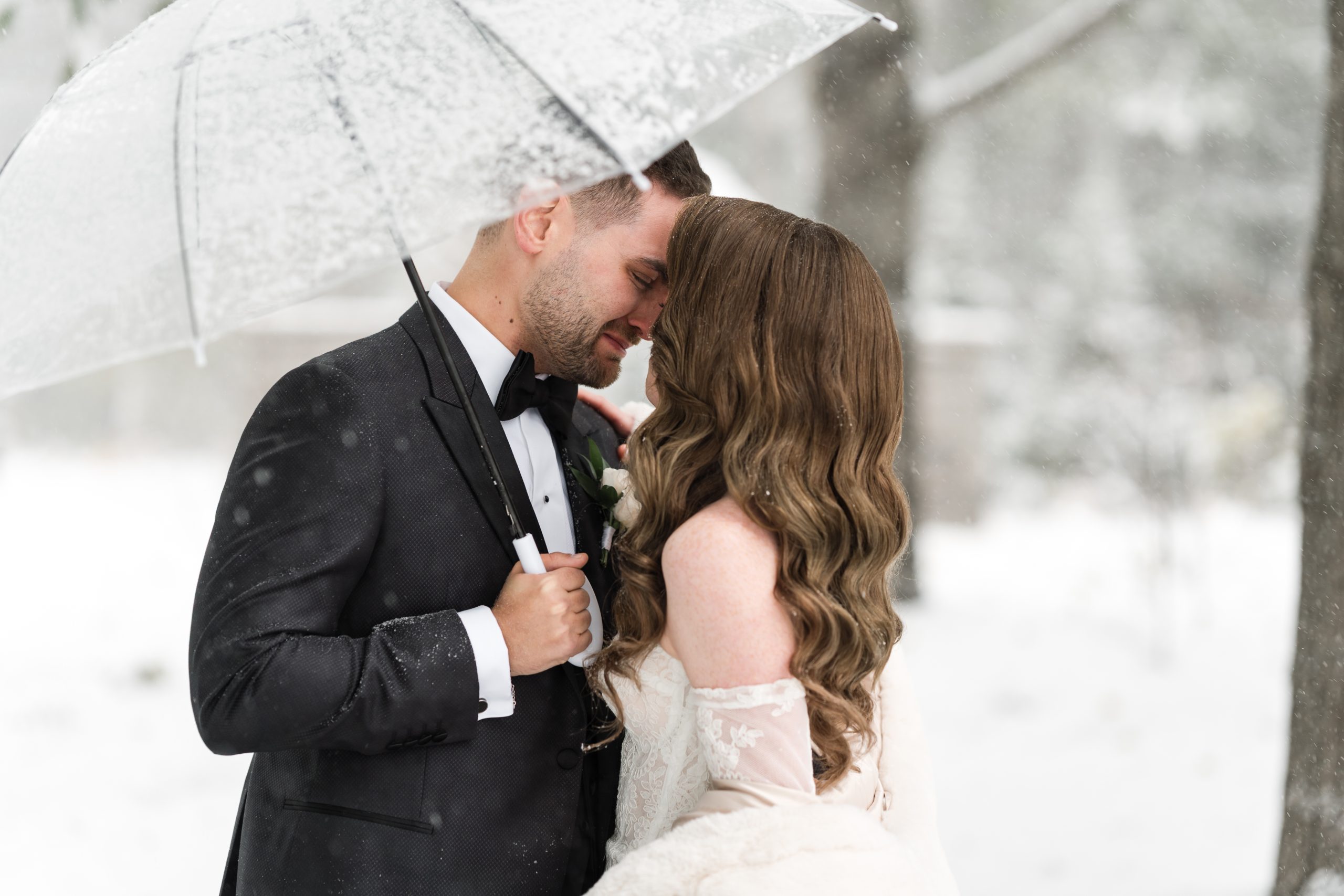 A couple dressed in formal wedding attire stands close together under a clear umbrella in a snowy outdoor setting, sharing an intimate moment. Snow falls gently around them, capturing the magic of winter photography in Toronto.