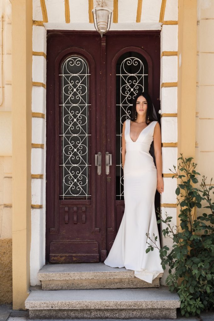 A woman in a white, sleeveless gown stands on steps in front of ornate double wooden doors with decorative ironwork, framed by a yellow and white building. A green plant grows beside her. Image taken by a Toronto wedding photographer