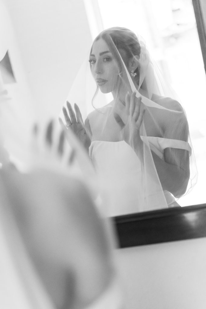 A bride wearing a veil and a white dress stands in front of a mirror, gently touching the glass with both hands and gazing at her reflection. The image is in black and white.