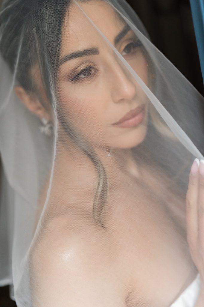 A bride with soft makeup and loose hair strands gazes thoughtfully while wearing a white veil over her face, capturing a serene and intimate moment before her wedding. Image taken by a Toronto wedding photographer