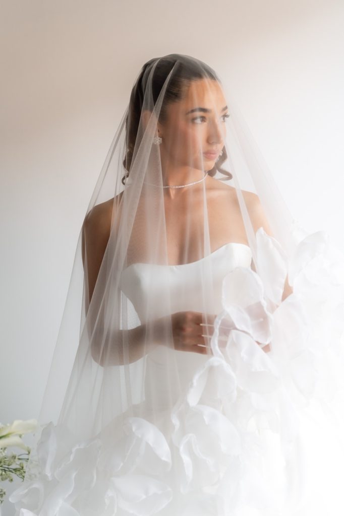 A bride in a strapless white wedding gown and veil holds a bouquet of white flowers, looking thoughtfully to the side against a softly lit, neutral background—captured beautifully by a Toronto wedding photographer.