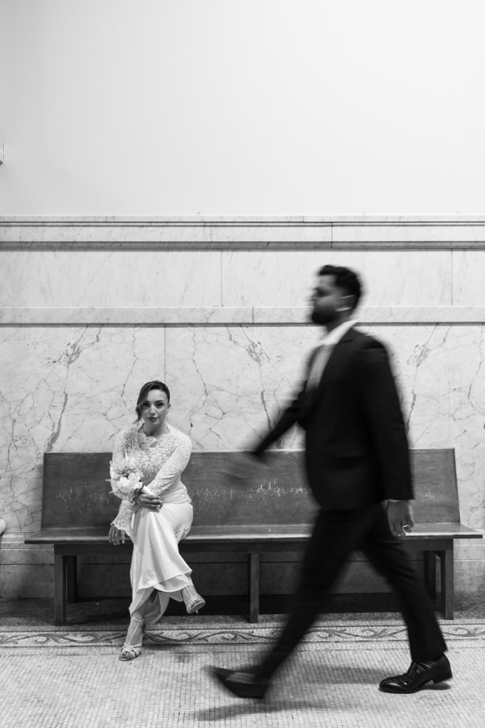 A bride in a white dress sits on a bench holding a bouquet, looking at the camera, while a man in a suit walks by in the foreground, slightly blurred. The scene is indoors with marble walls.