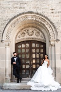 A bride in a white gown and a groom in a black tuxedo pose before an ornate, arched wooden doorway. Capturing timeless moments like these, Toronto wedding photography prices vary based on your unique style and setting.