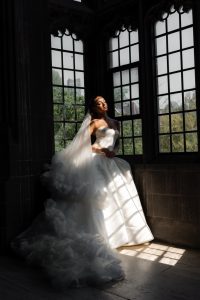 A bride in a strapless white wedding gown stands in sunlight by large window panes, her eyes closed and face tilted up, with a long, ruffled veil flowing behind her. Image taken at Hart House by a Toronto wedding photographer
