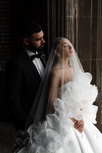 A bride in a white, ruffled gown and veil stands with her eyes closed, hands clasped, while a groom in a black tuxedo stands behind her, both framed by dark stone columns and soft natural light.