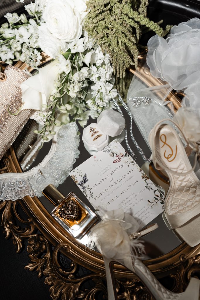 A decorative mirror displays wedding accessories: white flowers, lace garter, high heels, pearl jewelry, perfume, a clutch, and a wedding invitation with elegant gold script—perfectly styled for an editorial photographer capturing every enchanting detail.