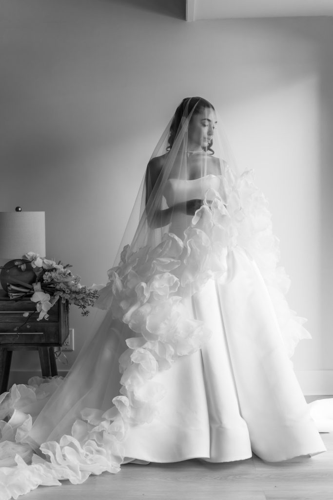 A bride in a voluminous, ruffled gown stands by a window in soft light, holding her dress. Captured by a Toronto photographer, the scene features a lamp and bouquet resting on a small table beside her. The photo is in black and white.