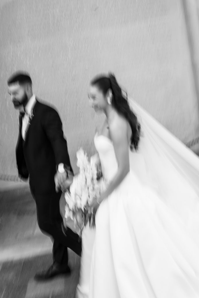 A blurred black-and-white photo by a Toronto wedding photographer captures a bride in a flowing gown holding flowers and a groom in a suit, holding hands as they walk together.