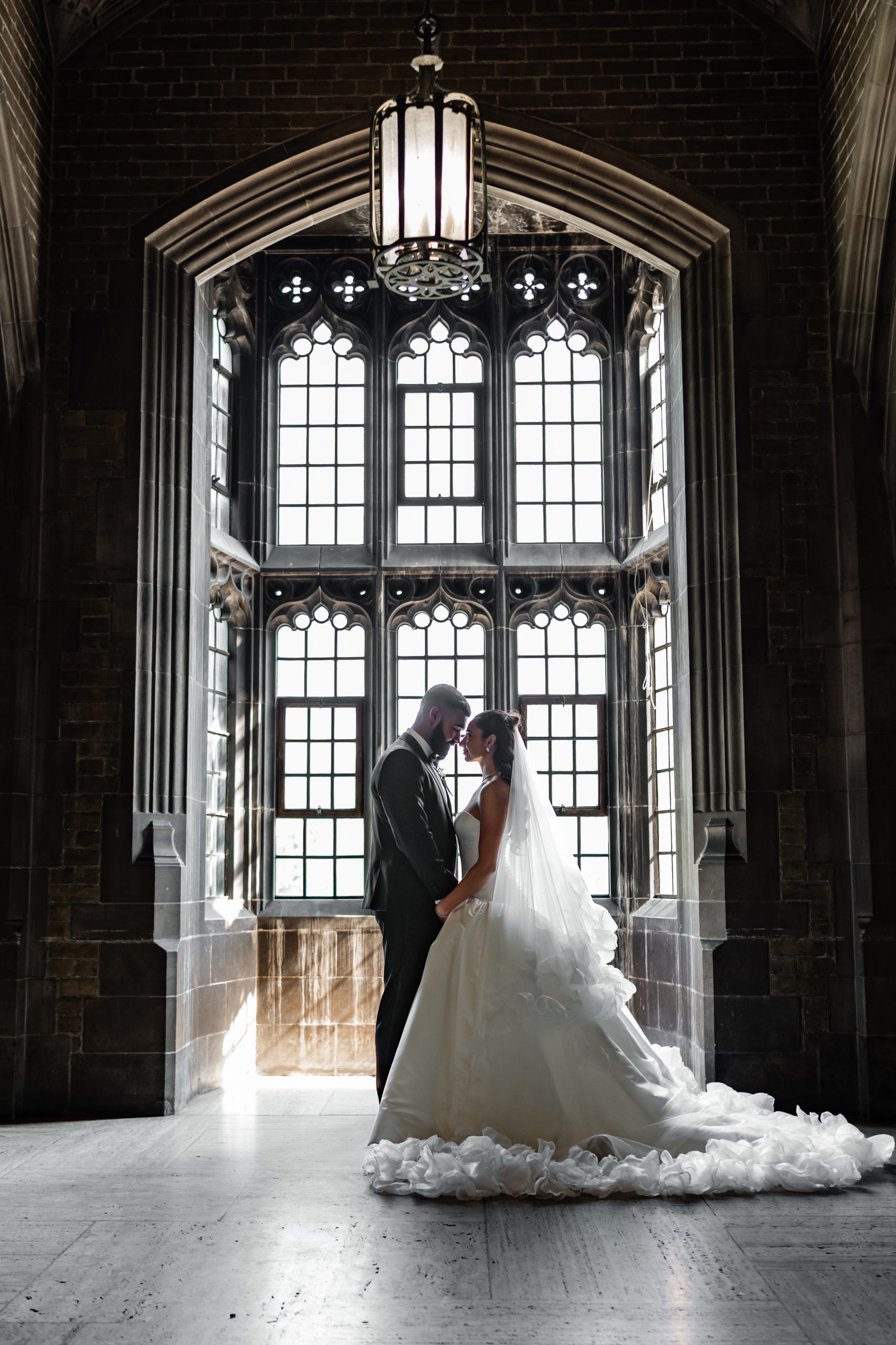 A bride and groom stand facing each other in front of a large, ornate window with gothic arches, softly lit by natural light. Captured in a documentary photography style, this intimate moment beautifully showcases the elegance of their Toronto wedding. The bride wears a long white gown and veil; the groom is in a dark suit.