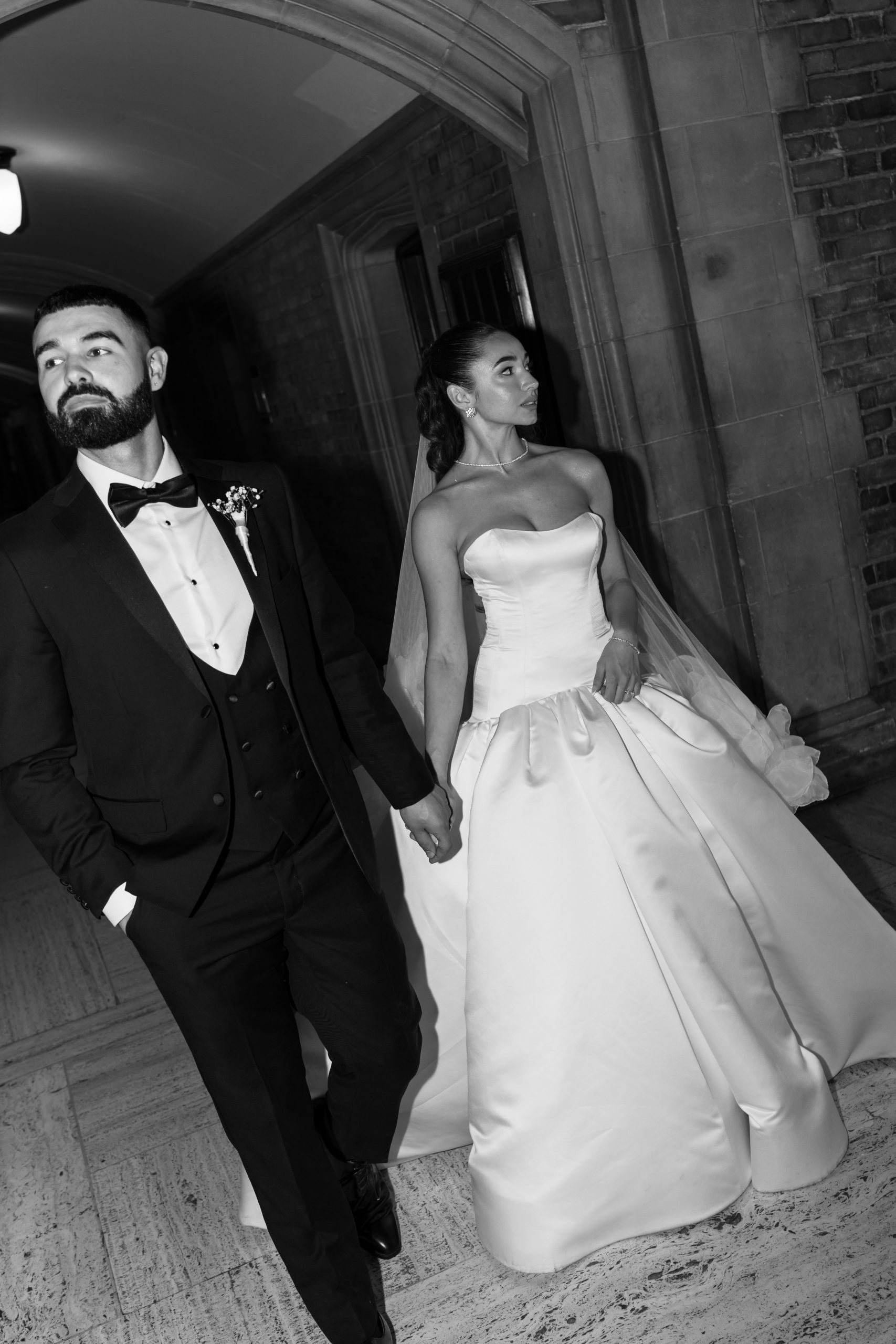 A bride in a strapless gown and a groom in a tuxedo hold hands while walking indoors, looking in different directions. This black and white photo captures a candid, documentary style that brings an editorial edge to the moment.