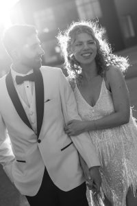 A couple dressed formally, the man in a white tuxedo and bow tie, and the woman in a sparkling dress, smile and hold hands outdoors in bright sunlight. The photo is in black and white.