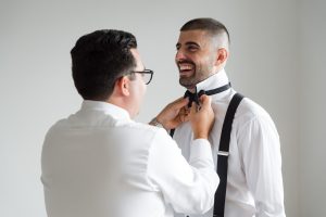 Two men in white shirts, one smiling as the other helps him adjust a black bow tie. Both appear happy, standing in a bright, plain room—captured in a candid, editorial style perfect for a wedding photographer’s portfolio.