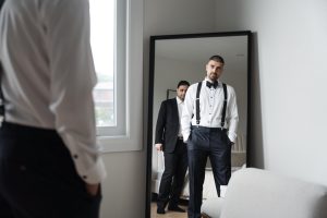 A man in formalwear with suspenders and a bow tie stands in front of a mirror, as a wedding photographer captures the moment; another man in a suit stands behind him. The room is modern with white walls and a window.