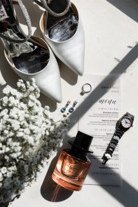 Editorial-inspired photography captures a flat lay of bridal accessories in Toronto: white heels, a menu, a silver watch, a ring, drop earrings, a perfume bottle, and delicate white baby's breath flowers, all artfully arranged on a sunlit white surface.