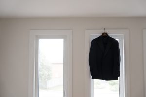 A black suit jacket hangs on a hanger between two tall, bright windows in a minimal, light-colored room, beautifully captured in wedding photography style.