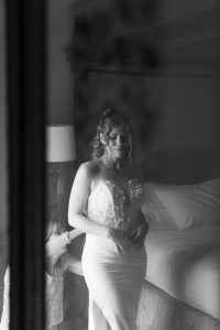 A bride in a strapless wedding gown stands near a bed, looking down, with another person adjusting her dress. The scene is softly lit and reflected in a mirror, capturing the timeless elegance favored by any Toronto photographer seeking an editorial touch.