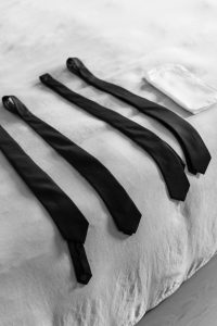 Four black neckties and a white pocket square are neatly laid out on a light-colored bedspread in black and white, creating an elegant scene perfect for wedding photography or a sophisticated editorial.